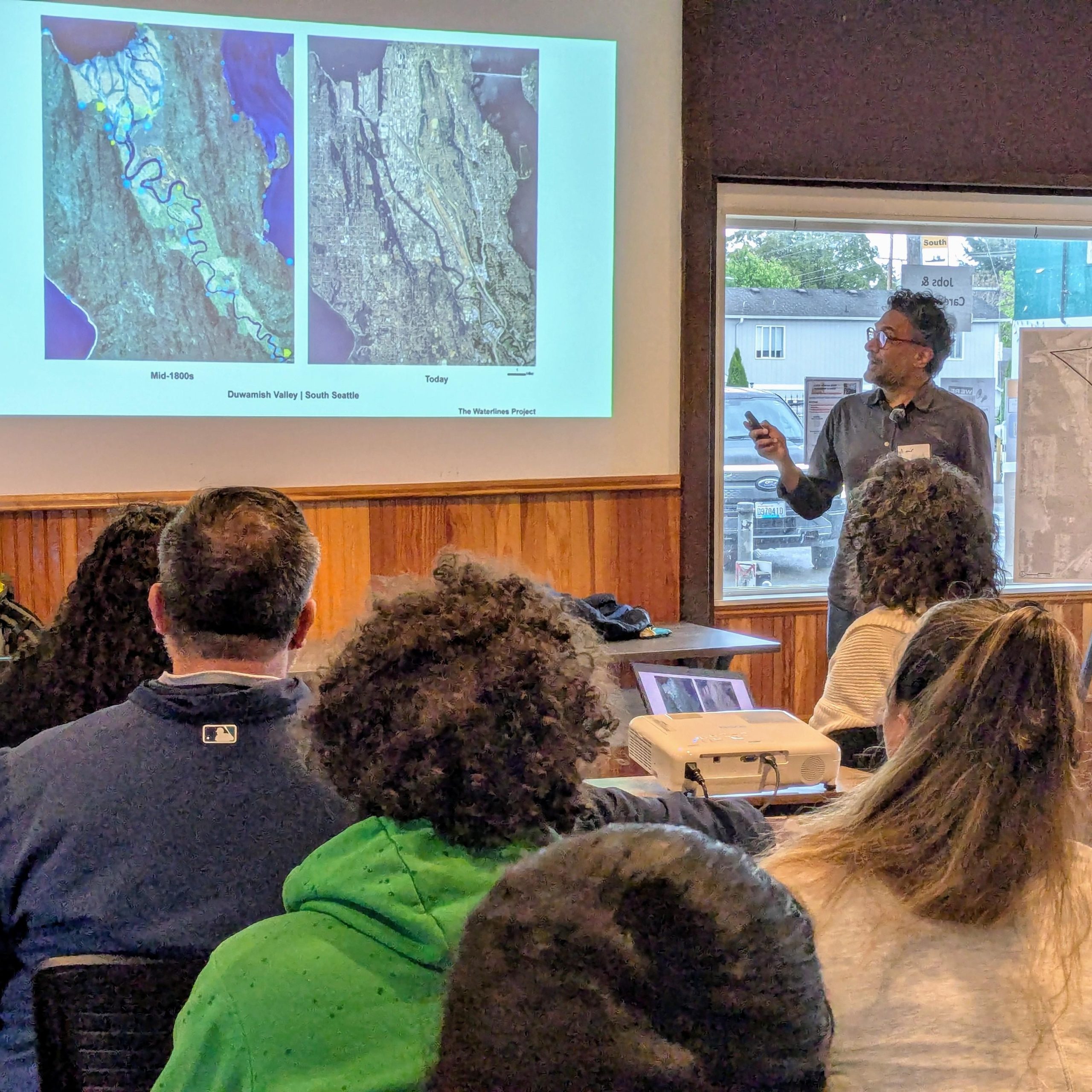 Man delivering a presentation to an audience, standing in front of a screen where two maps of the Duwamish Valley are projected