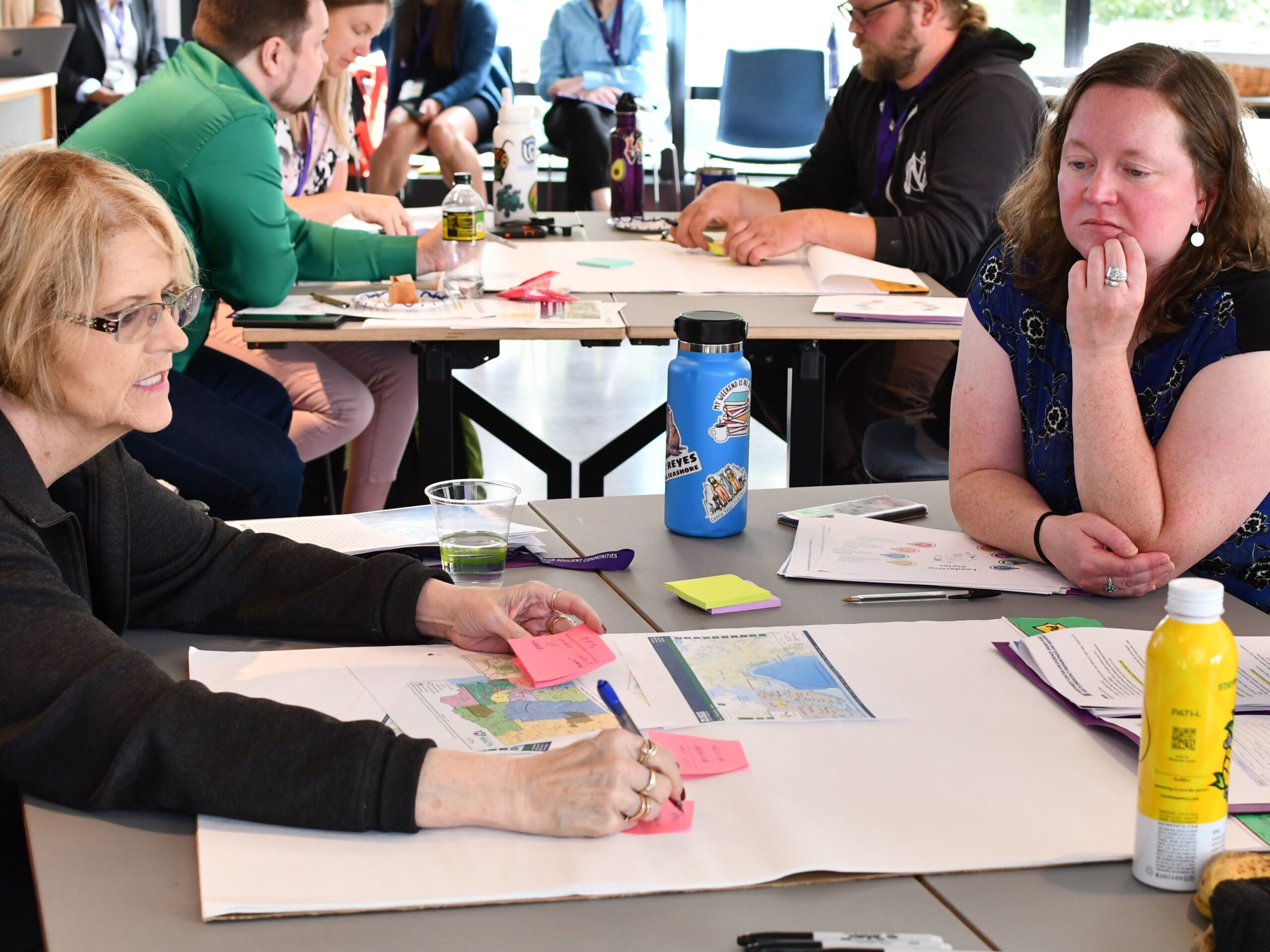 A woman talks to others seated at a table while she makes notes on a sheet of easel paper